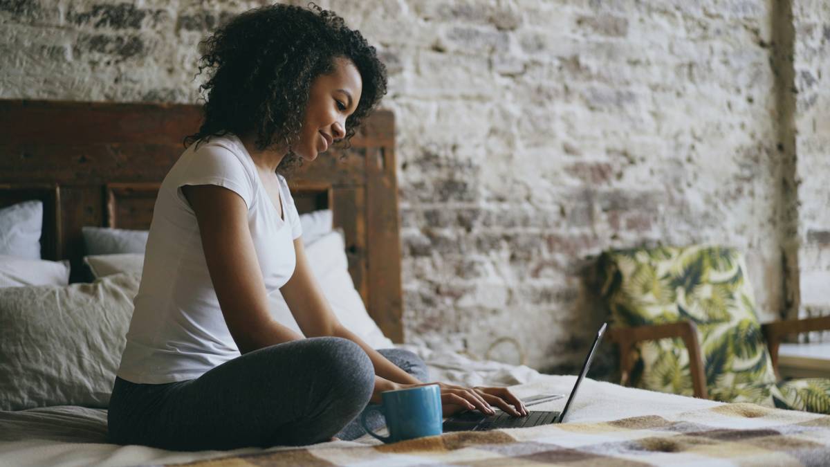 Cheerful mixed race girl typing on laptop computer for sharing social media sitting in bed and drinking coffee at home in the morning